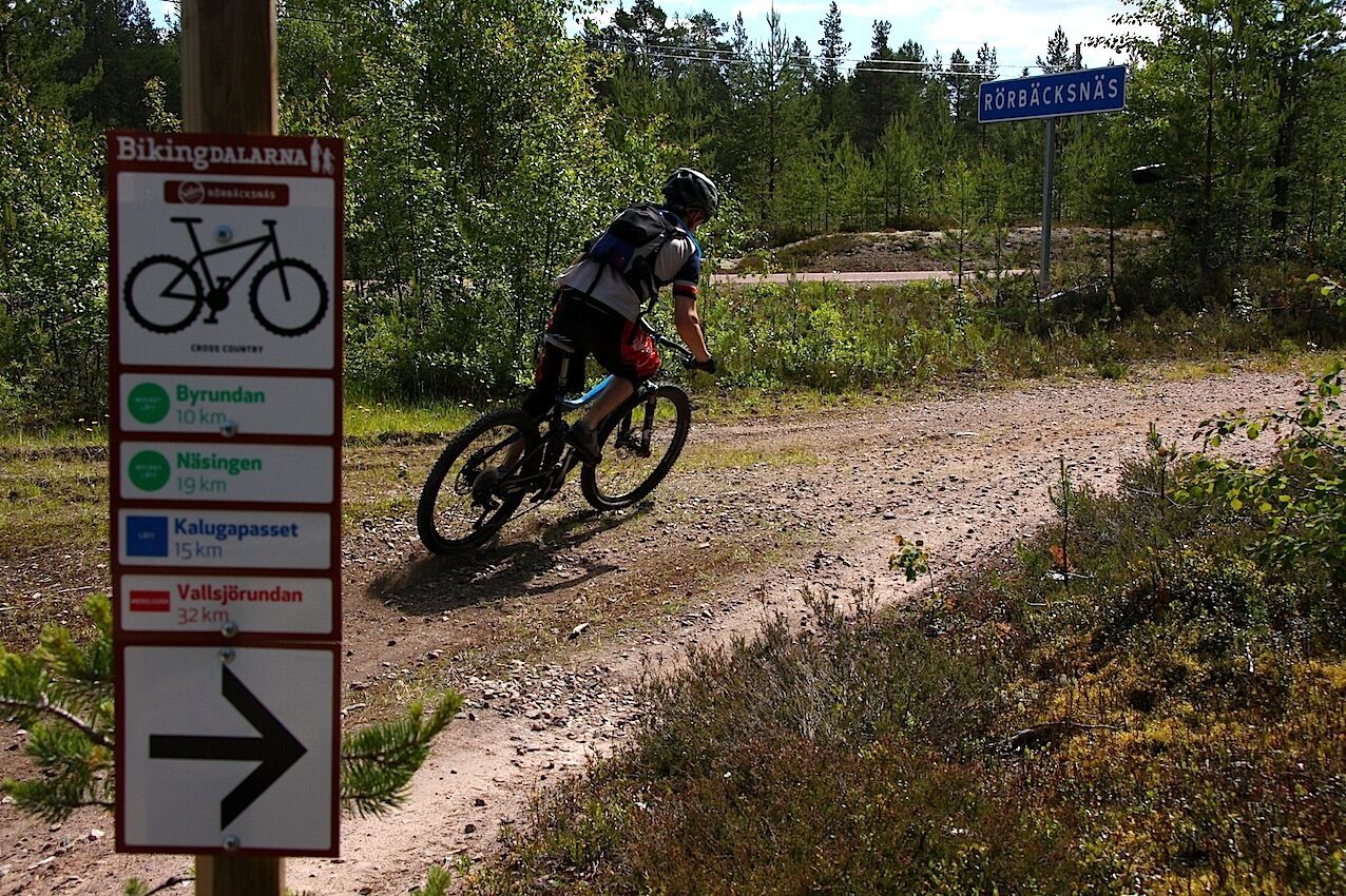 A mountain biker on a dirt trail with directional signs indicating distances to various locations.
