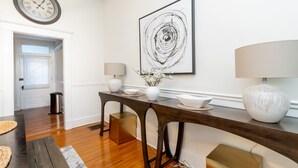 Un espace repas moderne avec une table en bois, deux chaises, un vase de fleurs, une horloge et un tableau abstrait encadré.