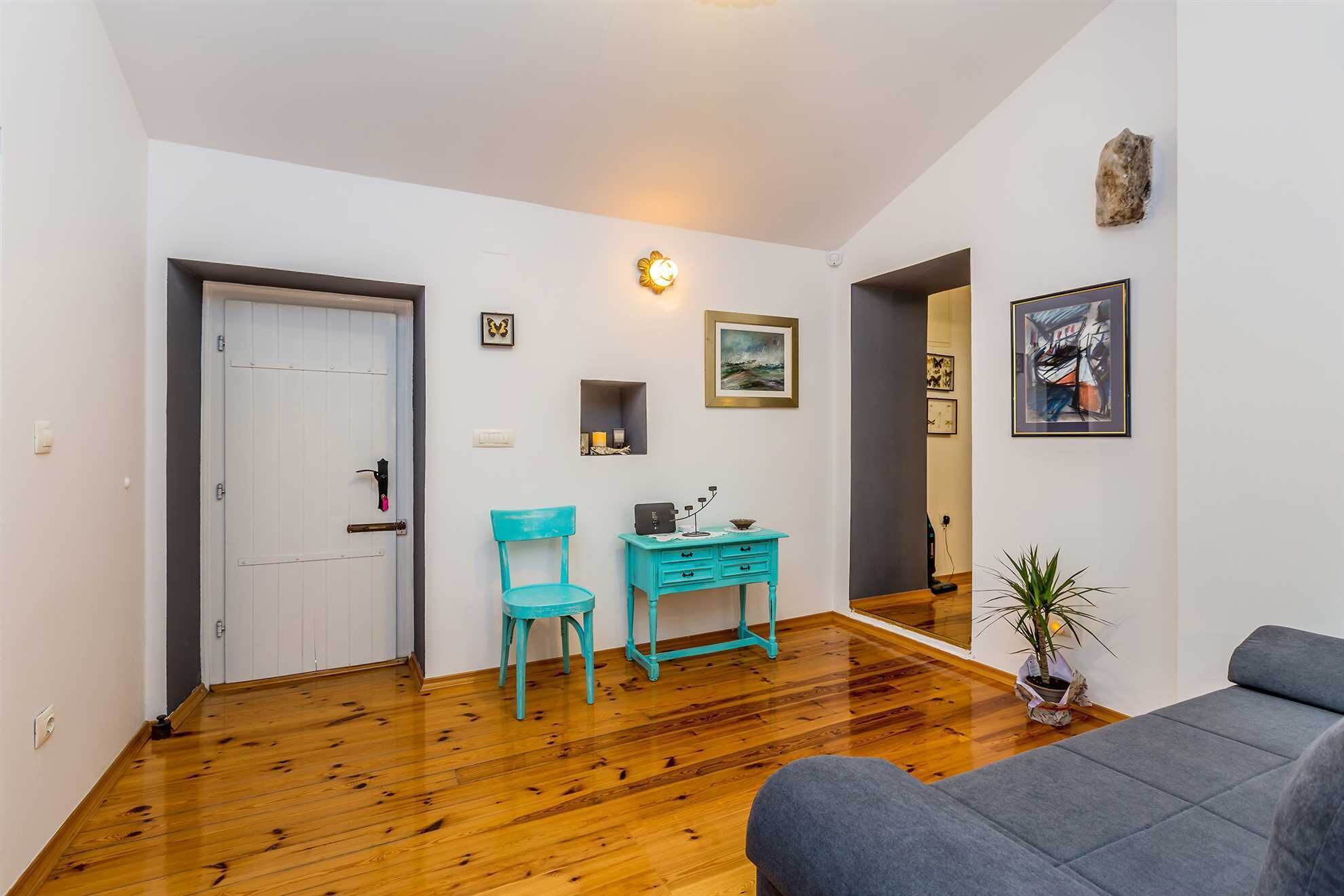 A room with a wooden floor, a white door, a turquoise chair, a small table with drawers, a grey sofa, and a potted plant.