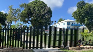 Une entrée fermée par un portail, agrémentée d’un panneau d’avertissement, un chemin menant à un bâtiment aux volets bleus et un jardin bien entretenu.
