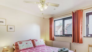 Ein Schlafzimmer mit Bett, Nachttisch, Fenster mit Vorhängen, Heizkörper und Deckenventilator.