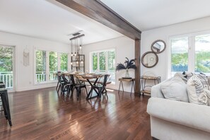 Amplia habitación con suelo de madera, zona de comedor con mesa y sillas de madera, sofá blanco y ventanas grandes que ofrecen vistas a zonas verdes.