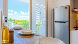 Una cocina moderna con un refrigerador de acero inoxidable, una mesa de comedor con platos y una botella, y vista a zonas verdes a través de un ventanal.