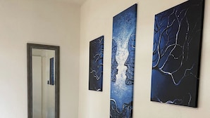A hallway with three abstract paintings on the wall, a mirror, and a bed with a patterned rug.
