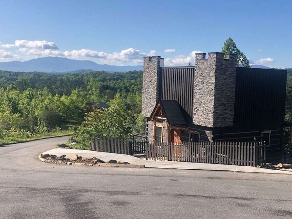 A stone building with a wooden door and a fenced driveway, surrounded by greenery and mountains.