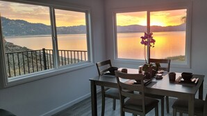 Un comedor con una mesa dispuesta para cuatro personas, dos ventanas con vistas a un lago y montañas, y un florero con flores en la mesa.