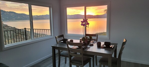 A dining area with a table set for four, two windows offering a view of a lake and mountains, and a vase with flowers on the table.