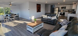 A modern living room with a leather sofa, dining table, and chairs. A kitchen with white cabinets and appliances is visible in the background.