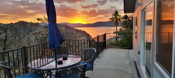 A balcony with a table and chairs, overlooking a body of water and mountains during sunset.
