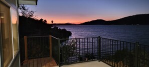 A balcony with a view of a calm lake at sunset, a wooden deck, and a metal railing.