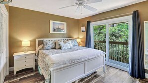 Un dormitorio con cama, mesitas de noche, ventilador de techo y vistas a la vegetación a través de un ventanal corredero.