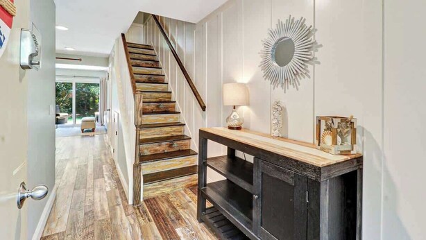 Una escalera de madera con peldaños que conducen a una plataforma, un espejo adosado a la pared y una consola con una lámpara y objetos decorativos.