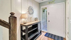 Un pasillo con escalera de madera, una consola con lámpara, un espejo decorativo y una puerta con la leyenda 'BEACH'.