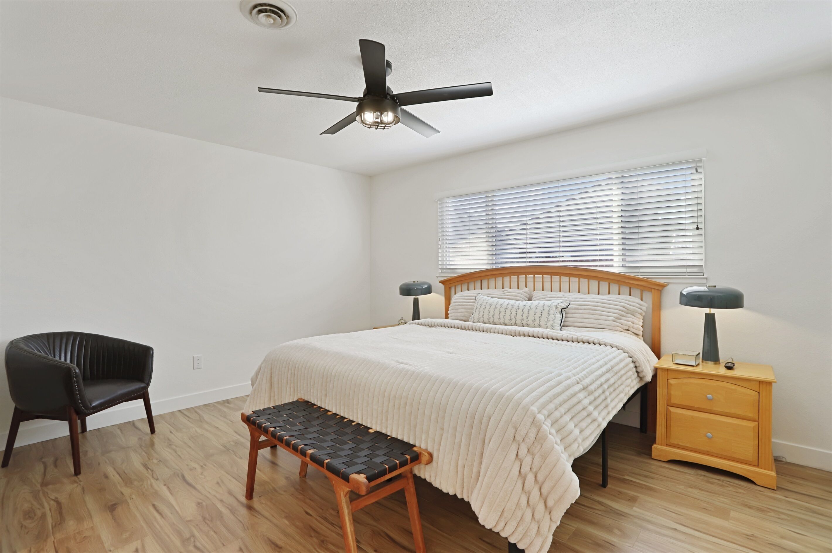 Ein Schlafzimmer mit einem Bett, einem Sessel, einer Bank, einem Nachttisch, einer Lampe, einem Deckenventilator und einem Fenster mit Jalousien.