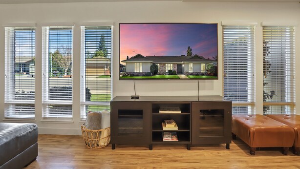 Un soggiorno con un'ampia televisione a schermo piatto montata a parete, un mobile porta TV in legno scuro, un divano grigio e un poggiapiedi in pelle marrone.