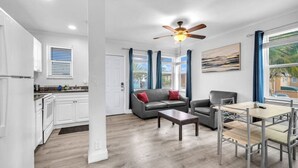 A modern living room with a grey sofa, a coffee table, and a dining area with a metal table and chairs. There is a ceiling fan, a window with blue curtains, and a painting on the wall. - Spectacular Ocean View Corner Apartment! (Hollywood)