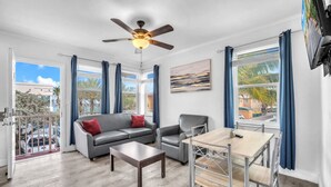 A spacious living room with a gray sofa, a wooden coffee table, and a dining table with chairs. There is a ceiling fan, large windows with blue curtains, and a painting on the wall. - Spectacular Ocean View Corner Apartment! (Hollywood)