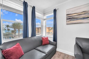 A modern living room with a grey sofa, red cushions, and a view of palm trees and buildings through large windows with blue curtains.