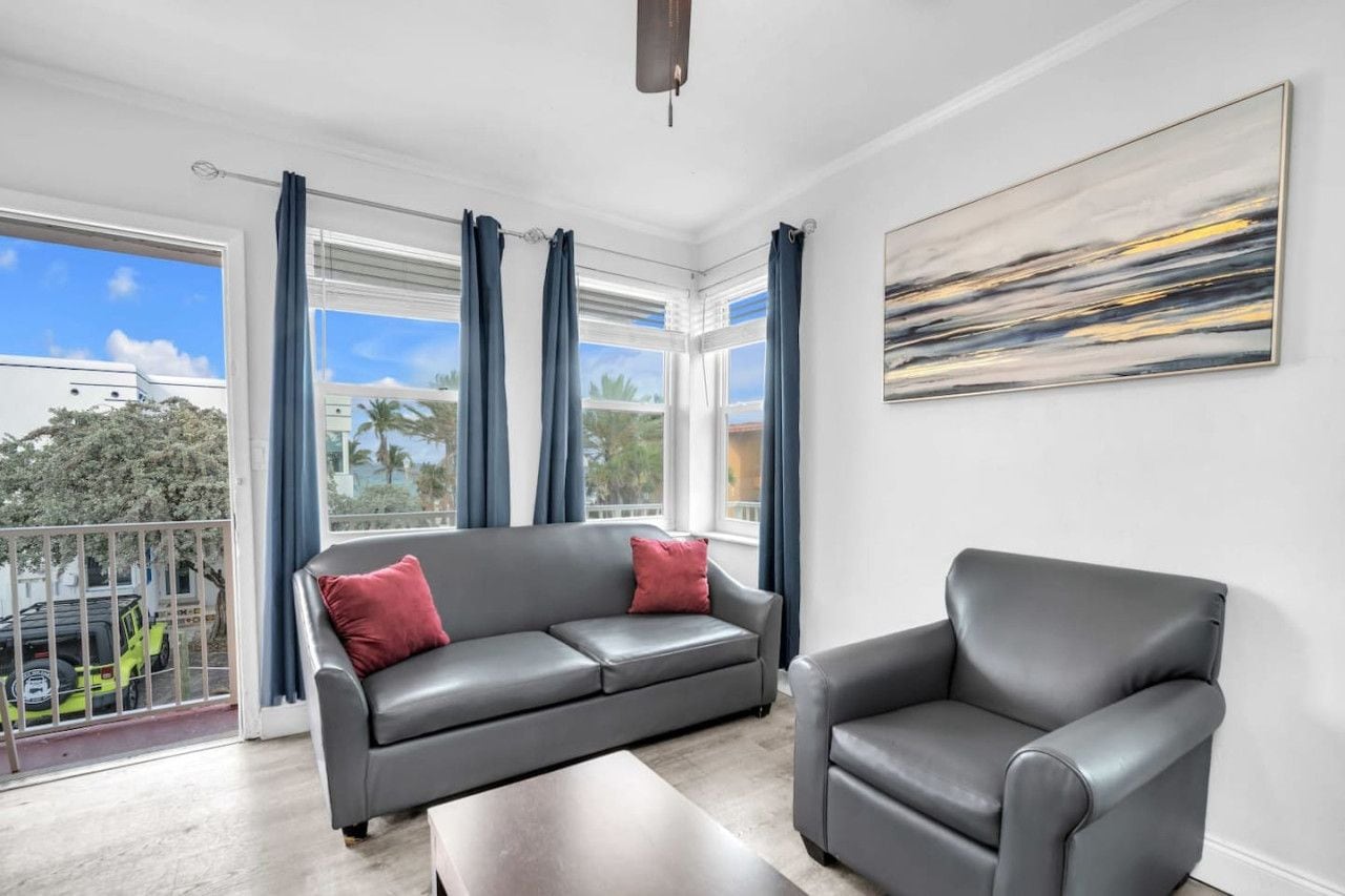 A modern living room with a grey sofa and armchair, large windows with curtains, and a painting on the wall.
