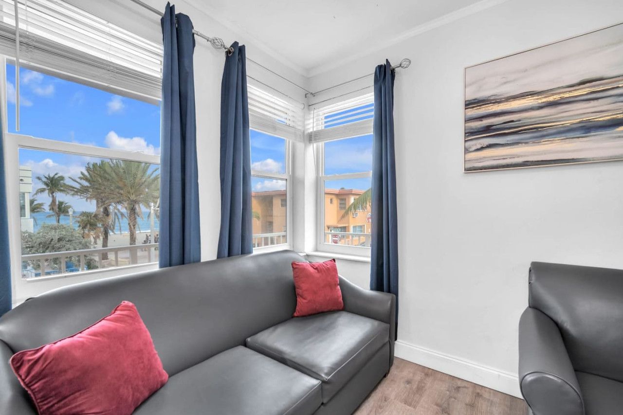 A modern living room with a grey sofa, red cushions, and a view of palm trees and buildings through large windows with blue curtains.