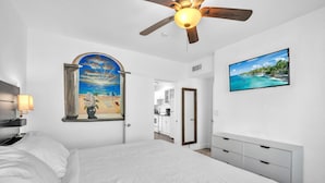 Een slaapkamer met een bed, een plafondventilator, een schilderij van een strand en een televisie aan de muur.