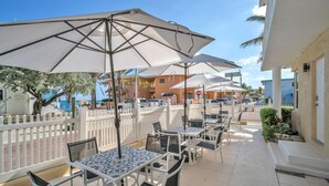 Een terras buiten met tafels en parasols, een wit hek en gebouwen op de achtergrond.