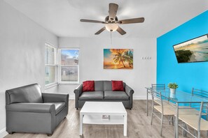 A modern living room with a grey sofa, a white coffee table, and a ceiling fan. There is a painting of a sunset on the wall and a TV mounted on the blue wall.