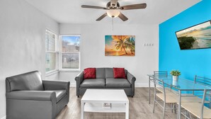 Ein modernes Wohnzimmer mit grauem Sofa, weißem Couchtisch und Deckenventilator. An der Wand hängt ein Gemälde von einem Sonnenuntergang und ein Fernseher ist an der blauen Wand angebracht.
