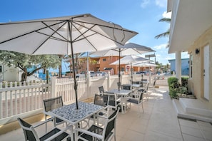An outdoor patio with tables and umbrellas, a white fence, and buildings in the background.