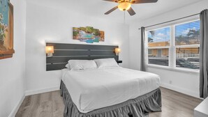 Un dormitorio con cama, ventilador de techo, mesita de noche, ventana con persianas y un cuadro en la pared.