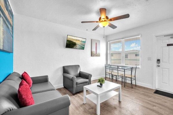 A modern living room with a grey sofa, a grey armchair, a white coffee table, and a ceiling fan. There is a dining table with chairs and a window with blinds.