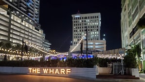 Een nachtzicht van een hotelentree met verlichte borden waarop 'THE WHARF' staat, en een modern gebouw op de achtergrond.