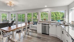 A spacious kitchen with white cabinets, a wooden dining table, and a view of a lush garden through large windows. - River Retreat W/ HOT TUB/ Game room (Bristol)