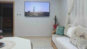 Un salón con un televisor de pantalla plana montado en la pared, un sofá con cojines estampados, una mesita con mantel blanco y una planta en maceta.
