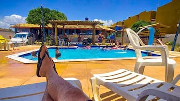 Zona de piscina con sillones de descanso, un área de estar techada y un edificio con tejado de tejas al fondo.