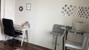 A modern living room with a black office chair, a grey sofa, and two bar stools. There is a small table with a 'Reserved' sign, a grey rug, and a wall decoration of hexagonal mirrors.