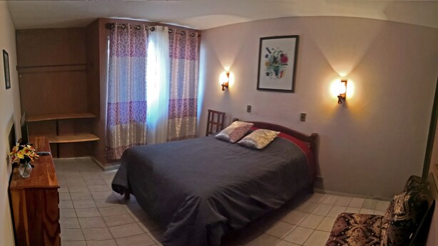 Un dormitorio con una cama, un aparador de madera, una silla y una lámpara fijada a la pared.