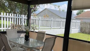 Eine überdachte Terrasse mit Tisch und Stühlen, einem Deckenventilator und Blick auf einen weißen Zaun sowie ein Haus im Hintergrund.