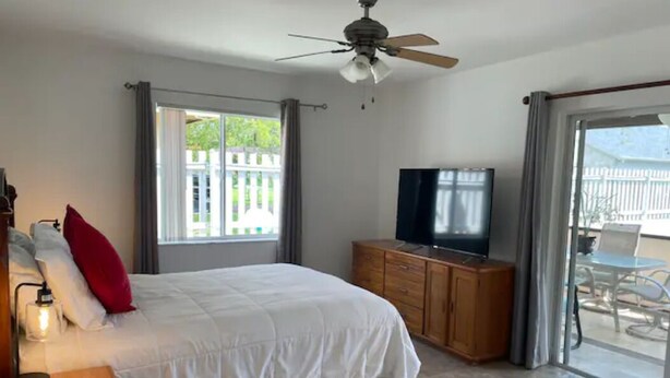 Ein Schlafzimmer mit Bett, Fernseher, Deckenventilator, Fenster mit Vorhängen und Blick ins Freie.