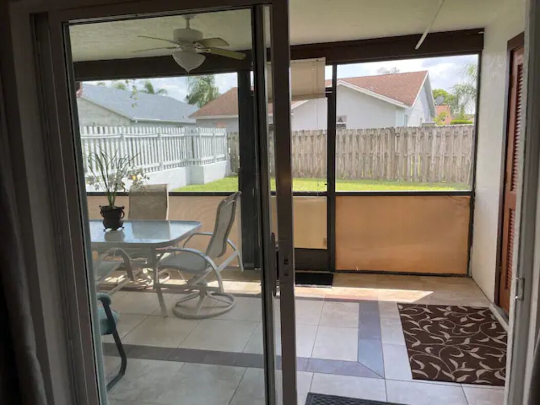 Une porte-fenêtre coulissante donne sur un patio aménagé avec une table et des chaises, un ventilateur de plafond et une vue sur un jardin avec une clôture en bois et une maison.