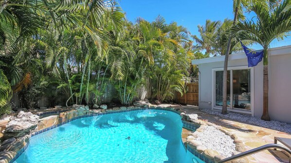 Une piscine entourée de plantes tropicales, une terrasse en pierre et un bâtiment moderne doté d’une porte en verre.
