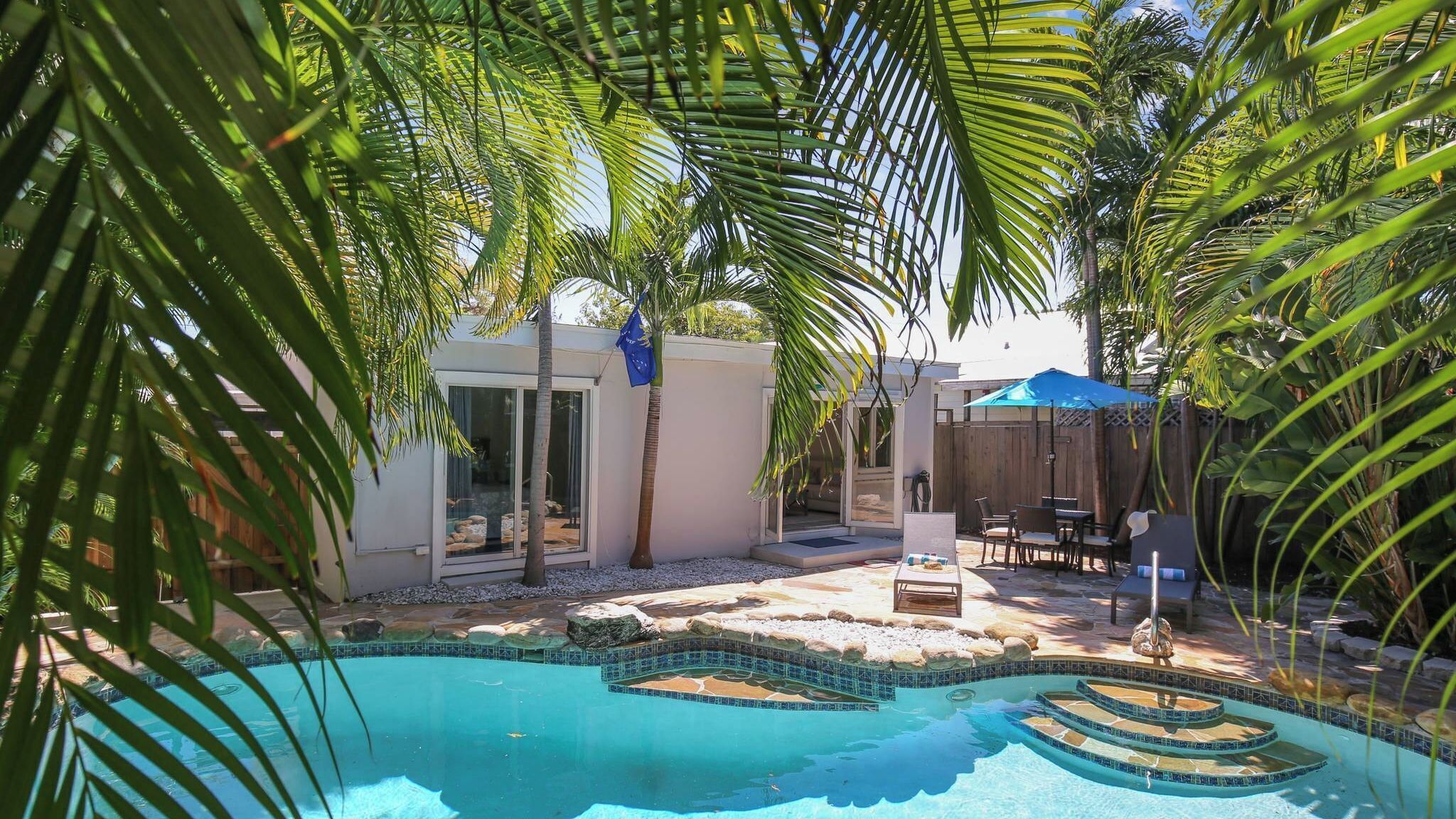 A backyard with a swimming pool, palm trees, and a patio area with outdoor furniture.