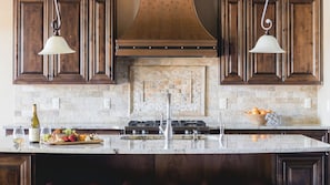 A kitchen with dark wooden cabinets, a stone backsplash, a central island with a sink, and pendant lights. - Klamath by Avantstay Premier Lakeside Cabin w/ Spa, Fire Pit & Lake Views (Bend)