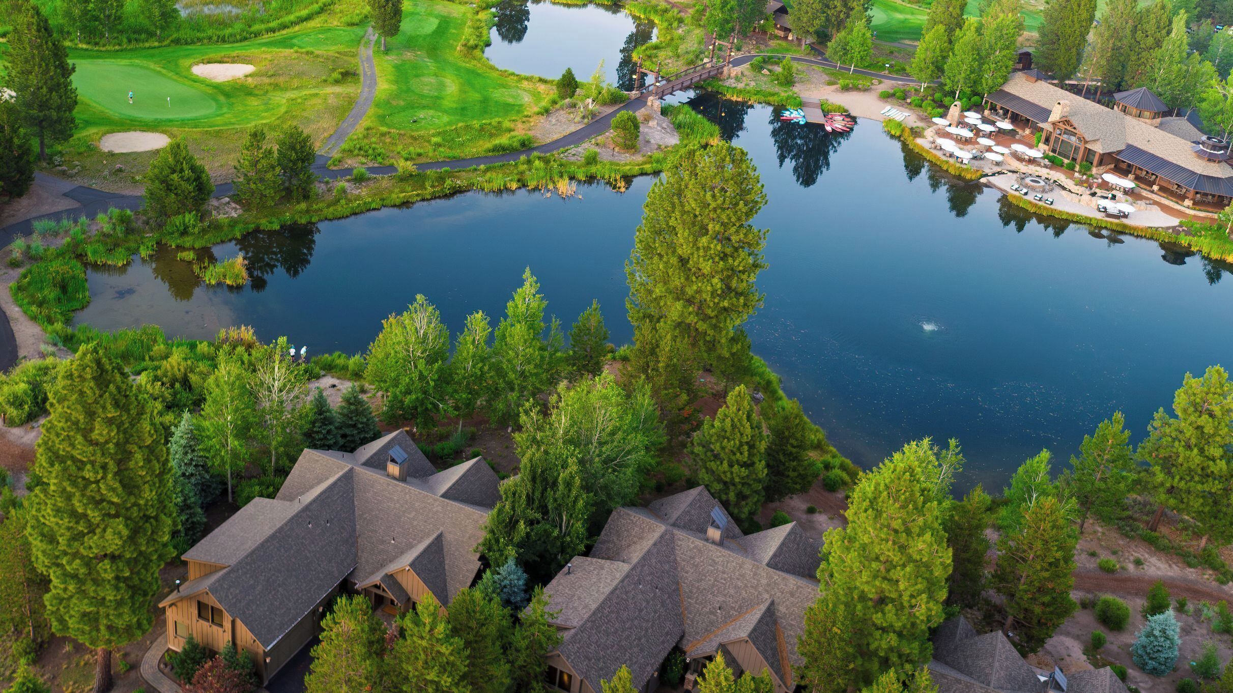 An aerial view of a golf course with a pond, surrounded by trees and buildings with sloped roofs.