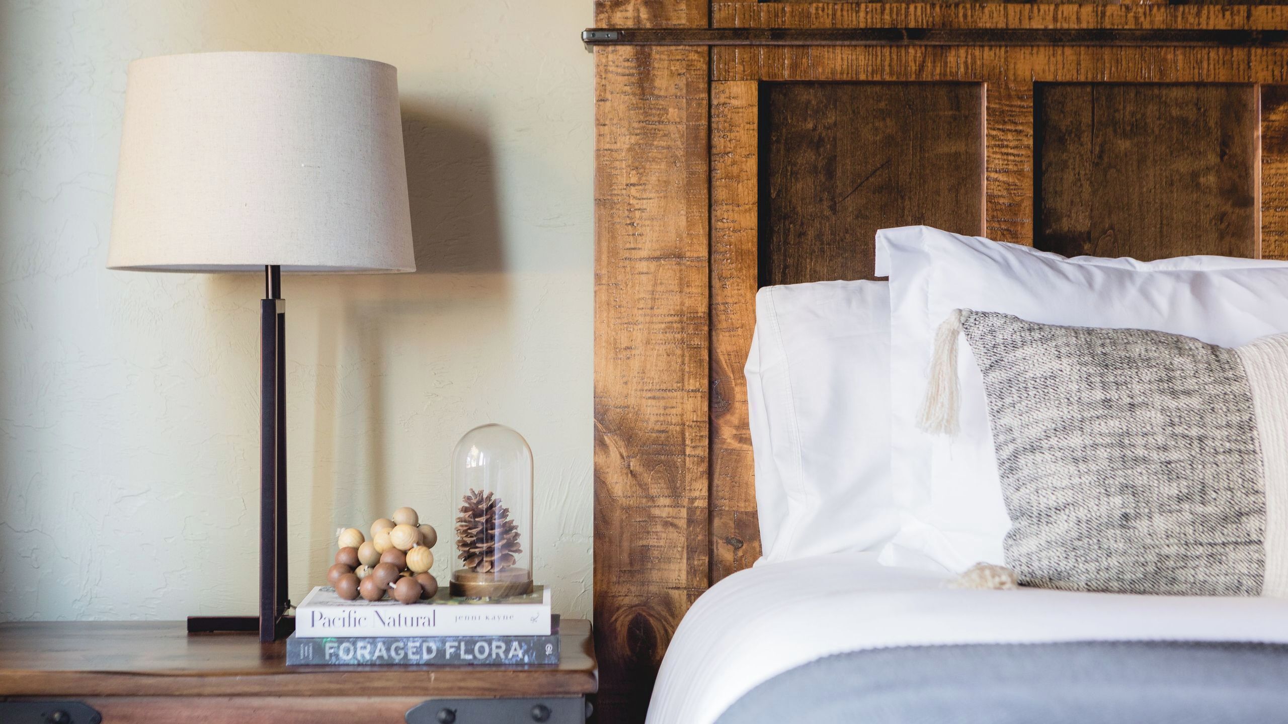A neatly made bed with white linens, a wooden headboard, a bedside lamp, and a small decorative arrangement including a book titled 'Pacific Natural' and a glass dome with pinecones.