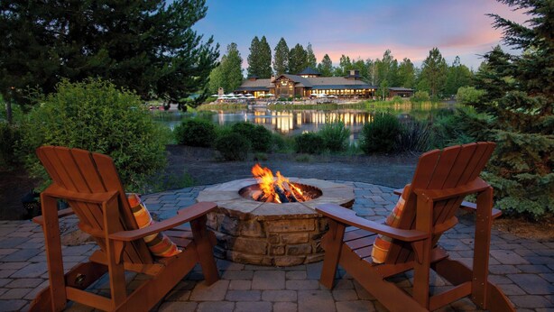 A fire pit with two Adirondack chairs, a pond, and a lodge in the background. - Klamath by Avantstay Premier Lakeside Cabin w/ Spa, Fire Pit & Lake Views (Bend)