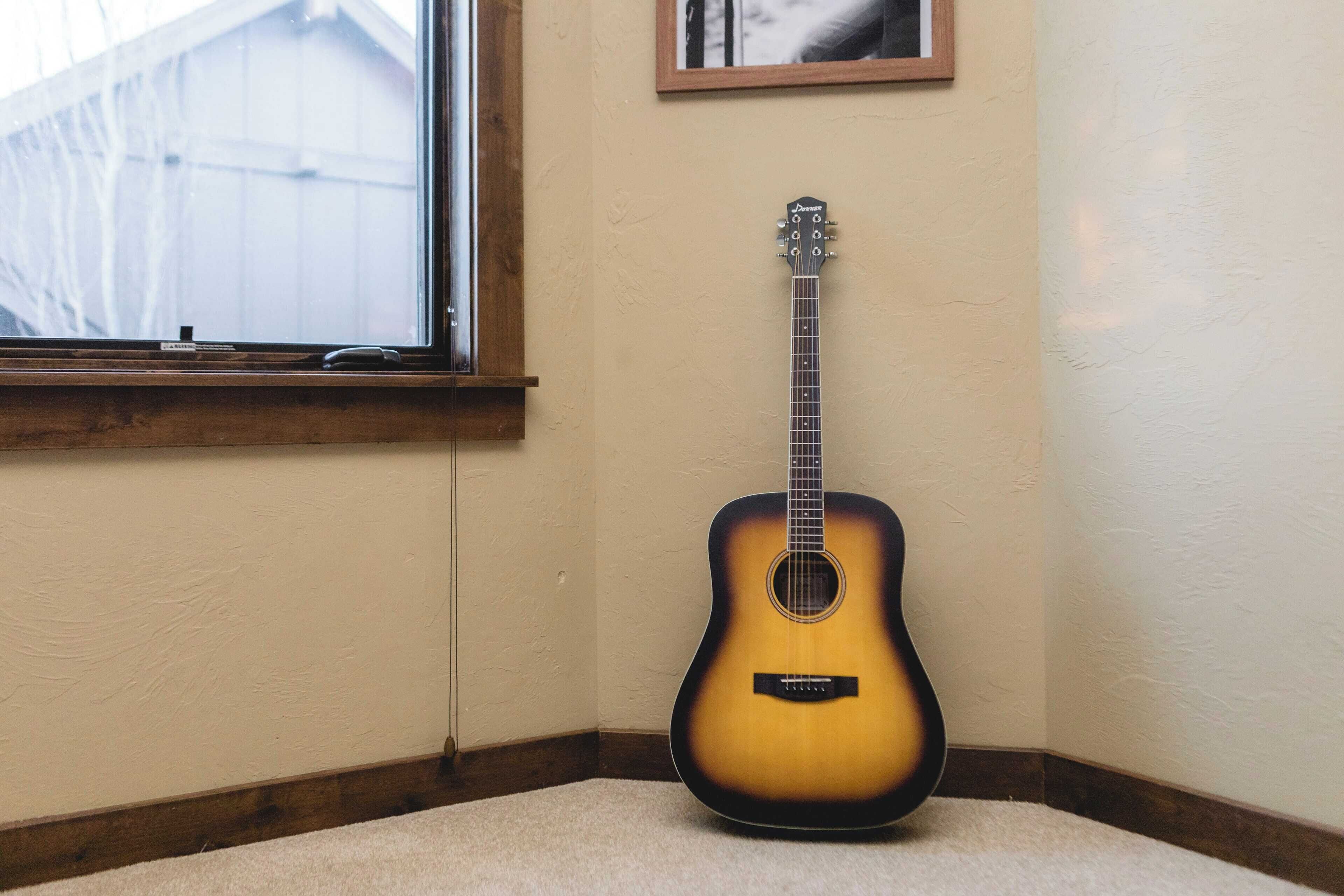 A guitar leaning against a wall in a corner with a window and a framed picture above it.