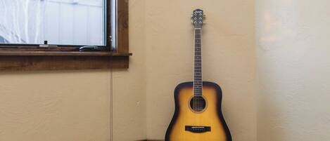A guitar leaning against a wall in a corner with a window and a framed picture above it.