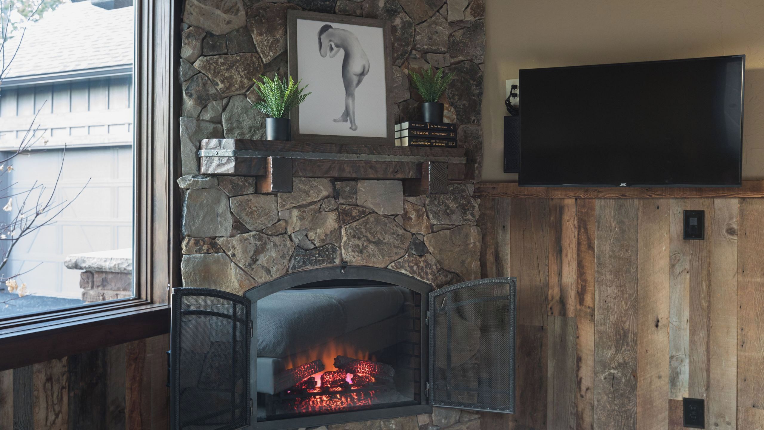 A stone fireplace with a fire, a framed artwork, and a television on a wooden wall.