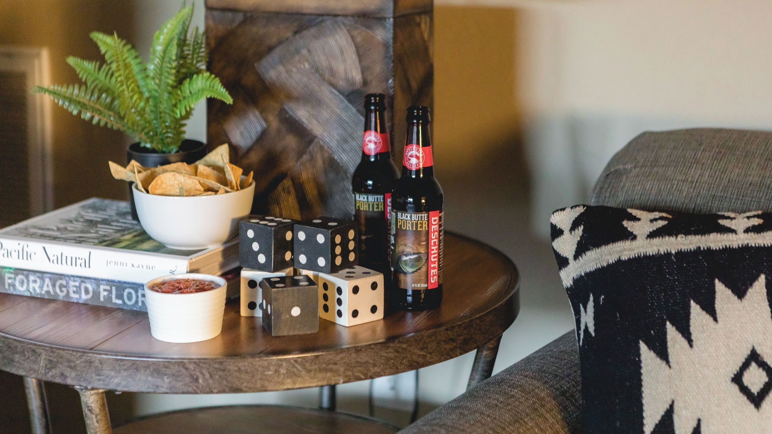 A table with a bowl of chips, two dice, a book titled 'Pacific Natural,' and two bottles of beer.